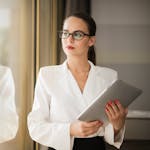 Businesswoman in formal attire thinking beside a window with a tablet.