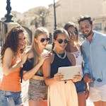 Five friends laughing together by the waterfront in Malta, enjoying a sunny day and sharing a tablet.