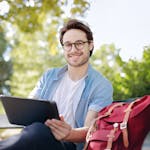 Smiling young man with glasses using a tablet on a park bench with a red backpack nearby.